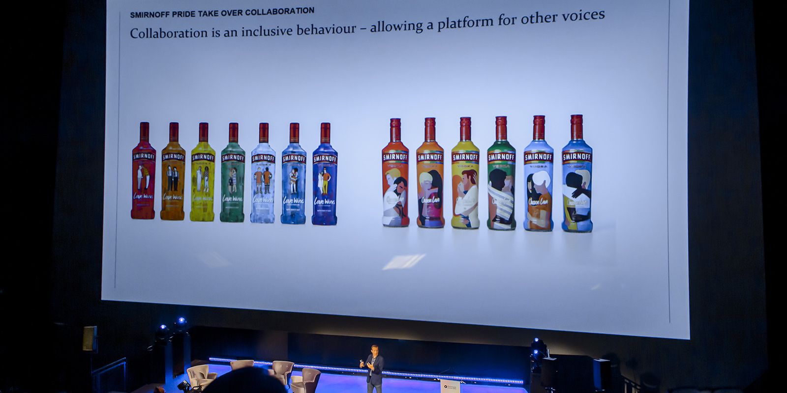 Jeremy Lindley en un escenario proyecta una diapositiva con botellas de vodka Smirnoff edición especial. Etiquetas ilustradas con parejas de diverso género y orientación sexual.