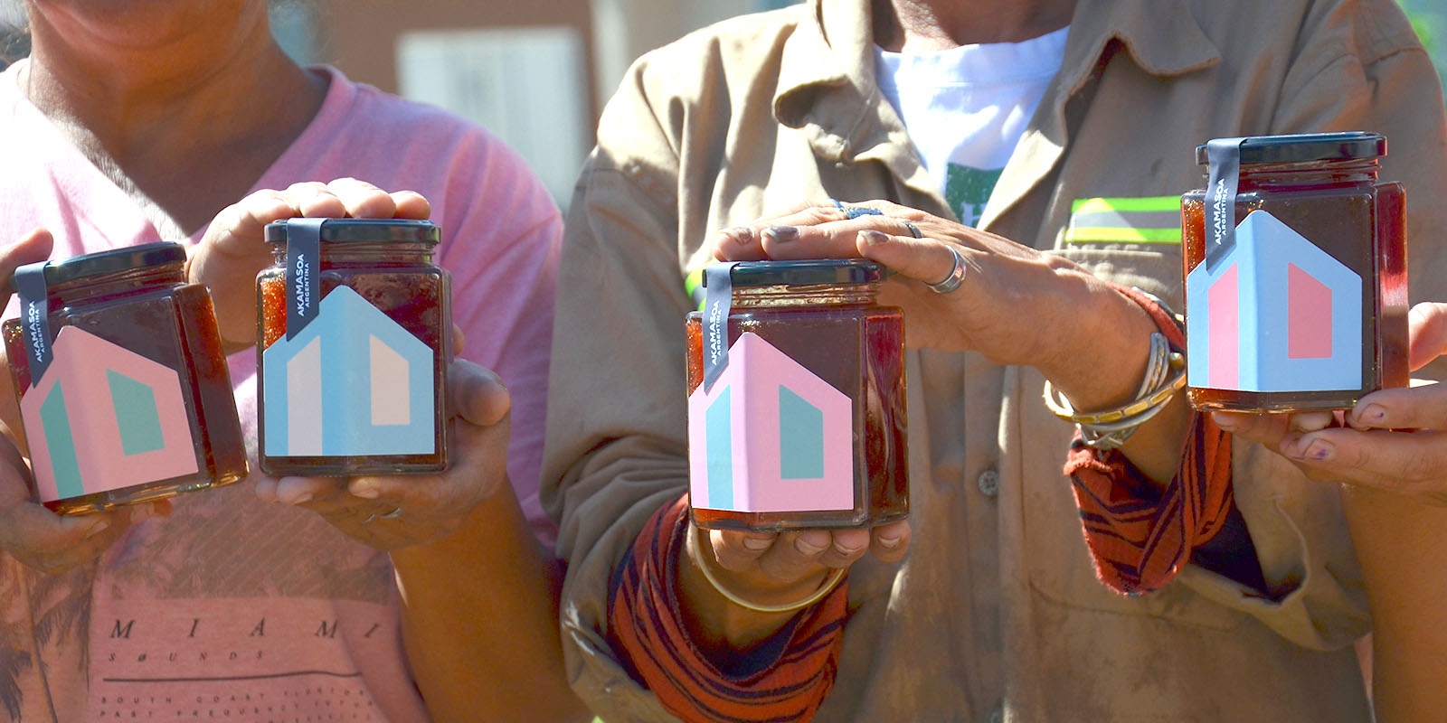 Mujeres trabajadoras de la comunidad Akamasoa Argentina mostrando frascos de mermelada con diseño por Tridimage, con etiquetas en colores rosa, azul y amarillo.