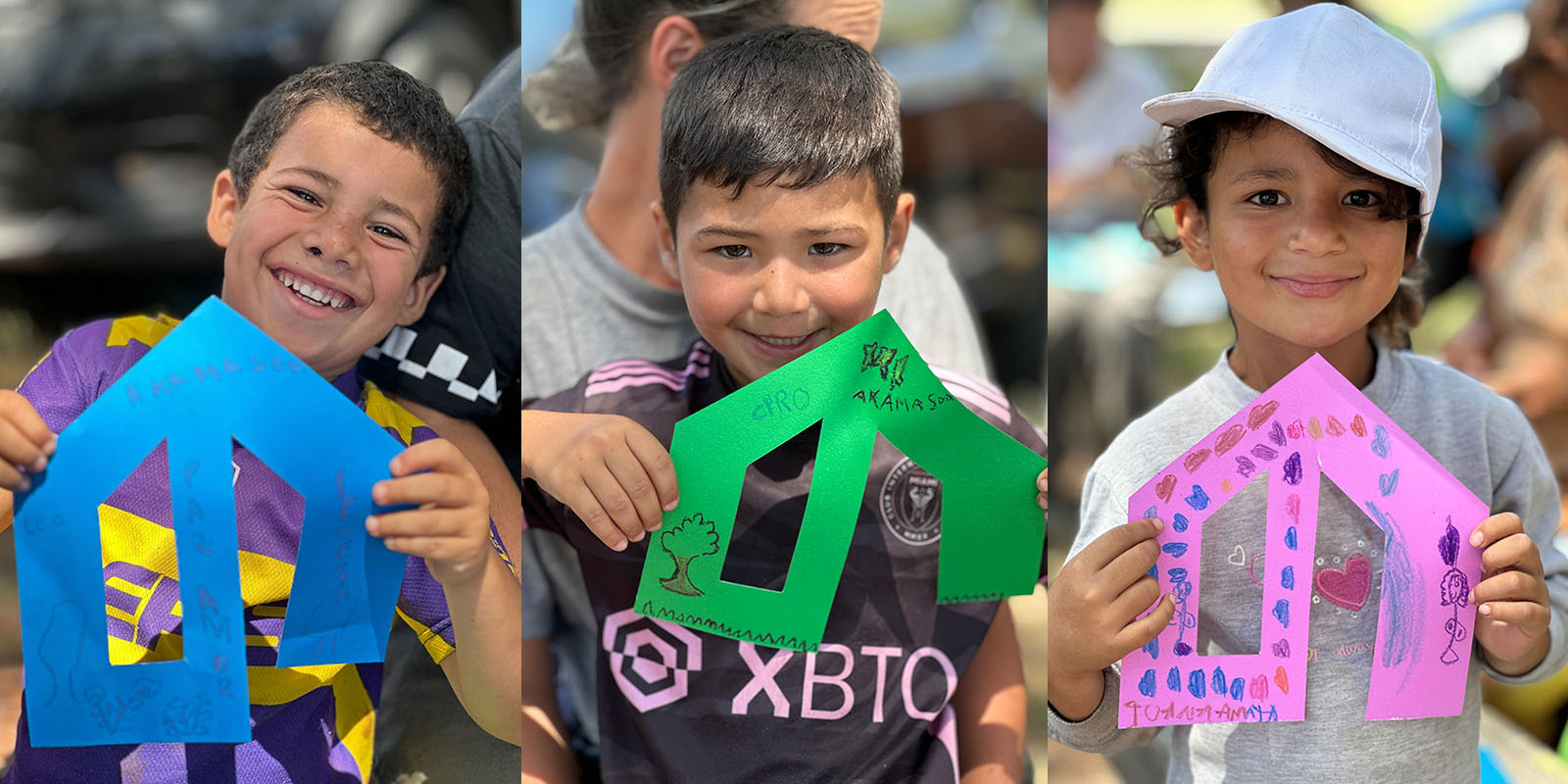 Dos niños y una niña humildes sonríen y muestran casas de cartulina dibujadas y coloreadas por ellos mismos, participando en una actividad creativa coordinada por Tridimage en Akamasoa Argentina.